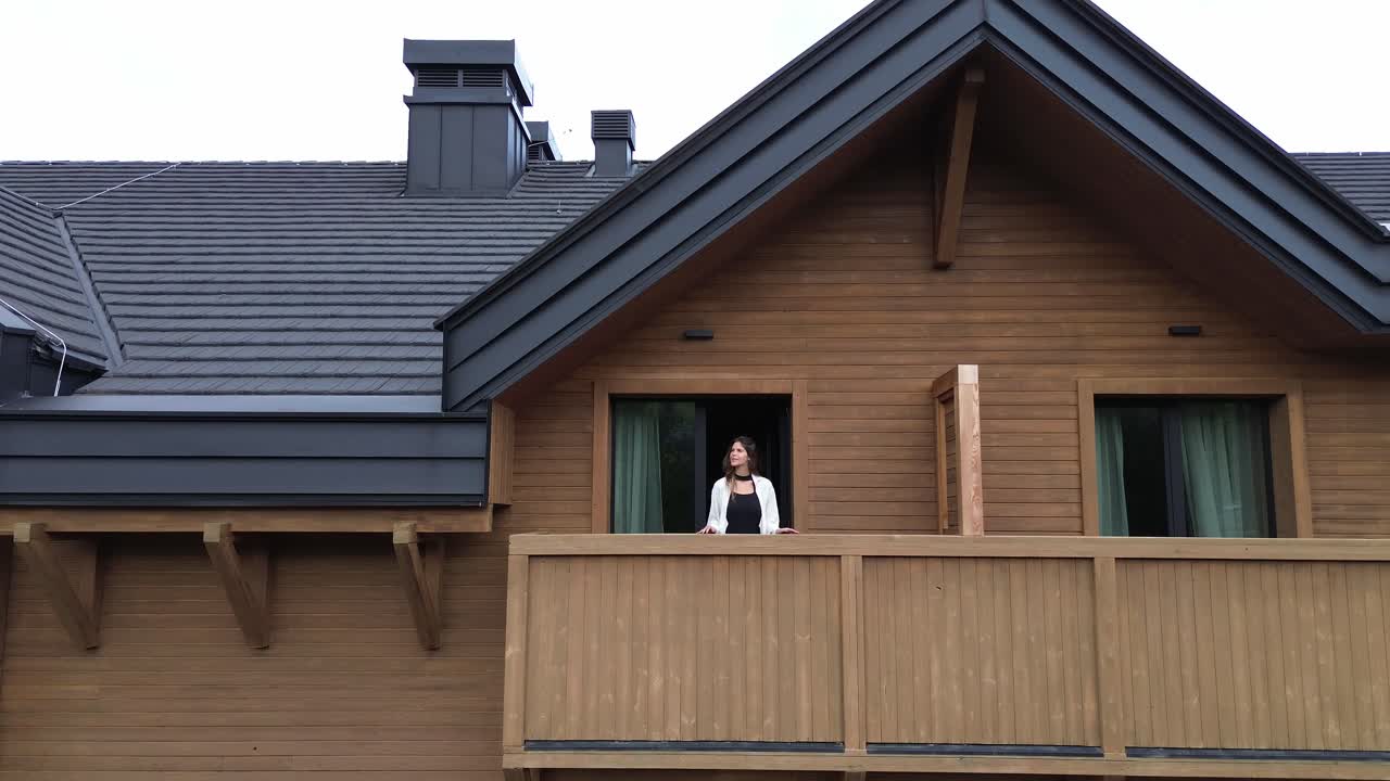 Woman standing on balcony of modern wooden chalet, Aerial pullback shot