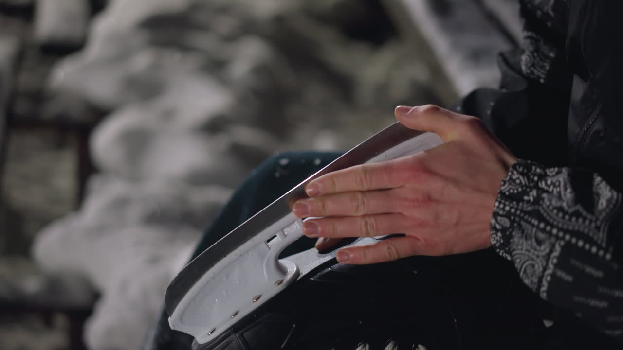 Close up of person holding and cleaning skating boot blade on snowy surface during cold winter night with soft lighting and background blur emphasizing hand motion and skate design details
