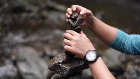 a close up of a man's hand wearing a watch stacking and balancing a rock on a river