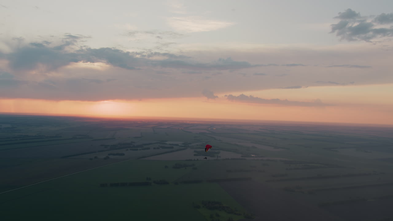 Distant silhouette of pilot gliding under red canopy across pastel sunset sky with soft clouds drifting over sprawling patchwork fields and tree lines, gentle breeze guiding serene evening