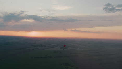 distant silhouette of pilot gliding under red canopy across pastel sunset sky with soft clouds drifting over sprawling patchwork fields and tree lines, gentle breeze guiding serene evening