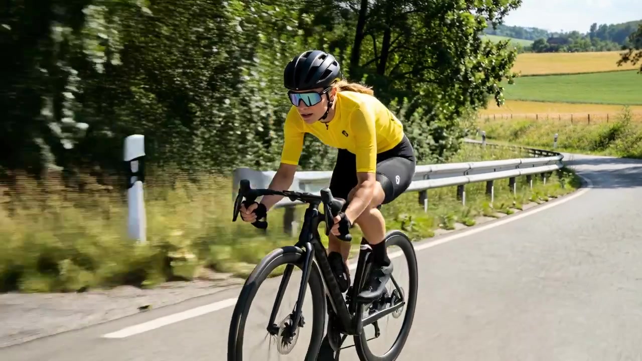 Ciclista in piega con maglia gialla che si avvicina a una curva a destra su una strada rurale, pedalando una bici in carbonio per l'equilibrio