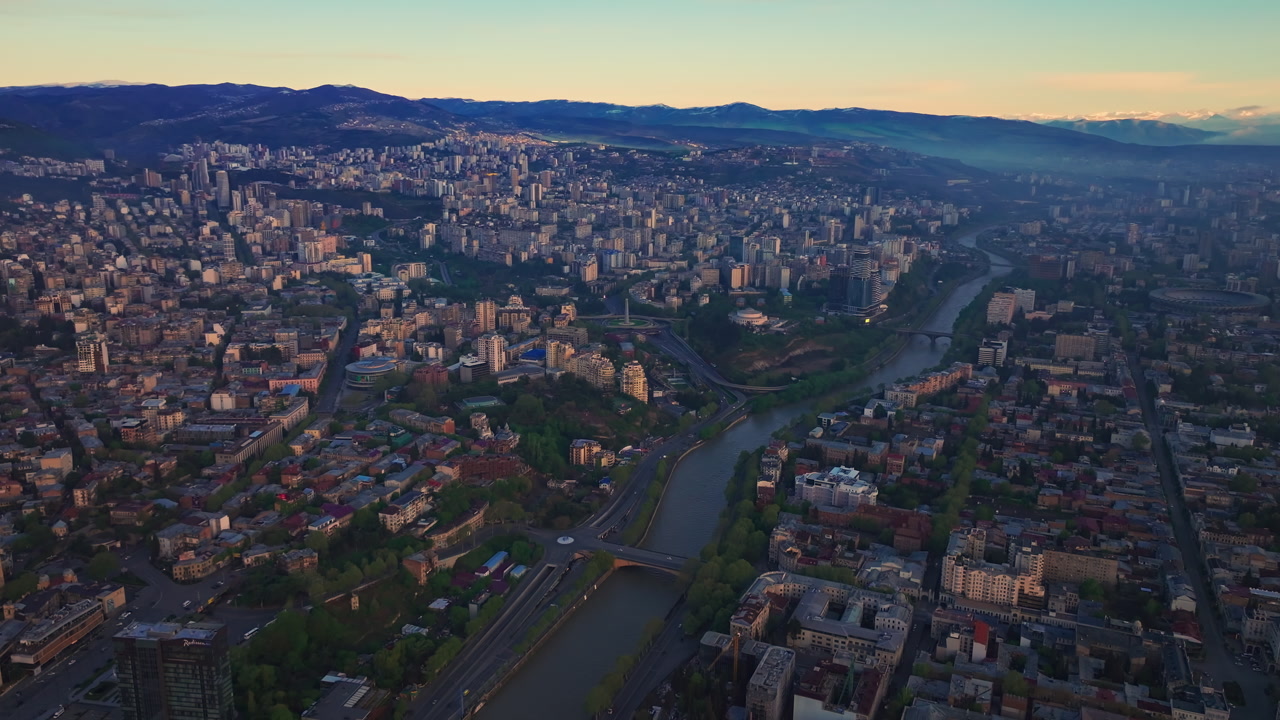 Aerial view of Tbilisi, Georgia, with the river winding through the city and mountains rising in the background, revealing urban texture, historic districts, and dramatic natural surroundings.
