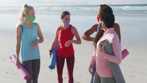 group of diverse female friends wearing face masks holding yoga mats at the beach touching elbows