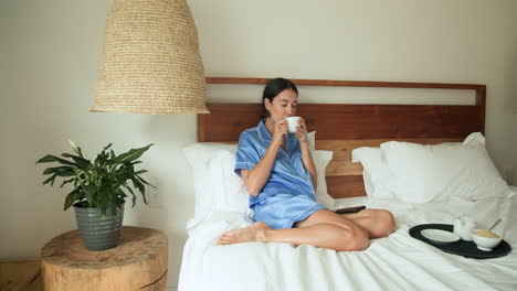 Relaxing in bed, woman in blue pajamas enjoying morning coffee, smiling peacefully, at home