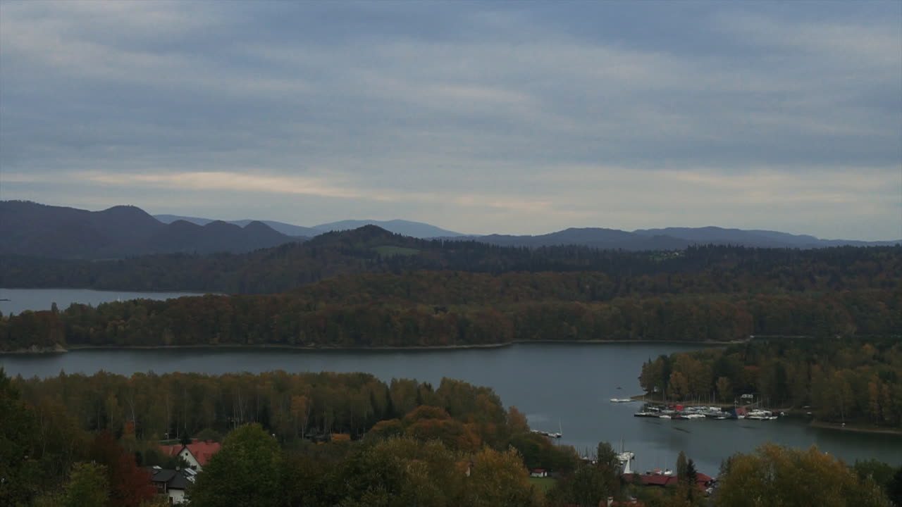 Timelapse af en stor sø mellem skovklædte bakker under en overskyet himmel. Sejlbåde lægger til ved bredden, fjerne bjerge forsvinder i tågen. Fredfyldt og malerisk landskabsbillede af Solina-søen, Polen.