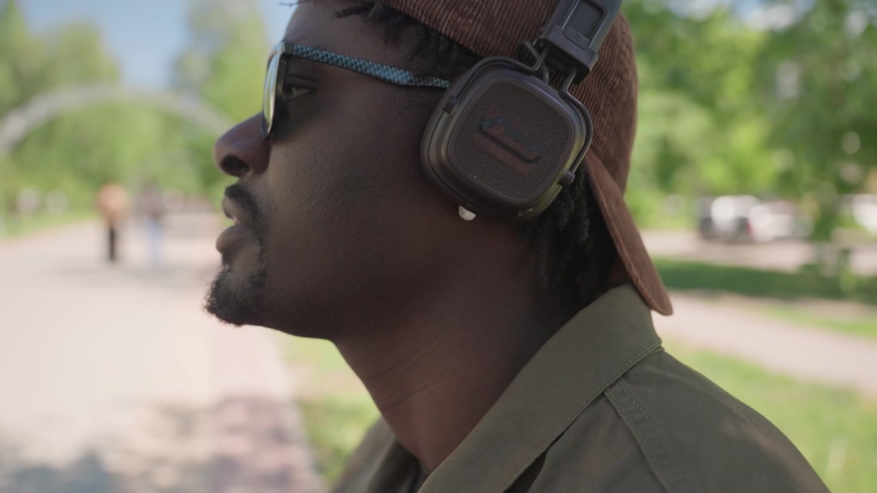 Close up of man wearing sunglasses and headphones standing outdoors with greenery in background. Headphones are black cap is brown corduroy shirt is olive drab. Soft natural light