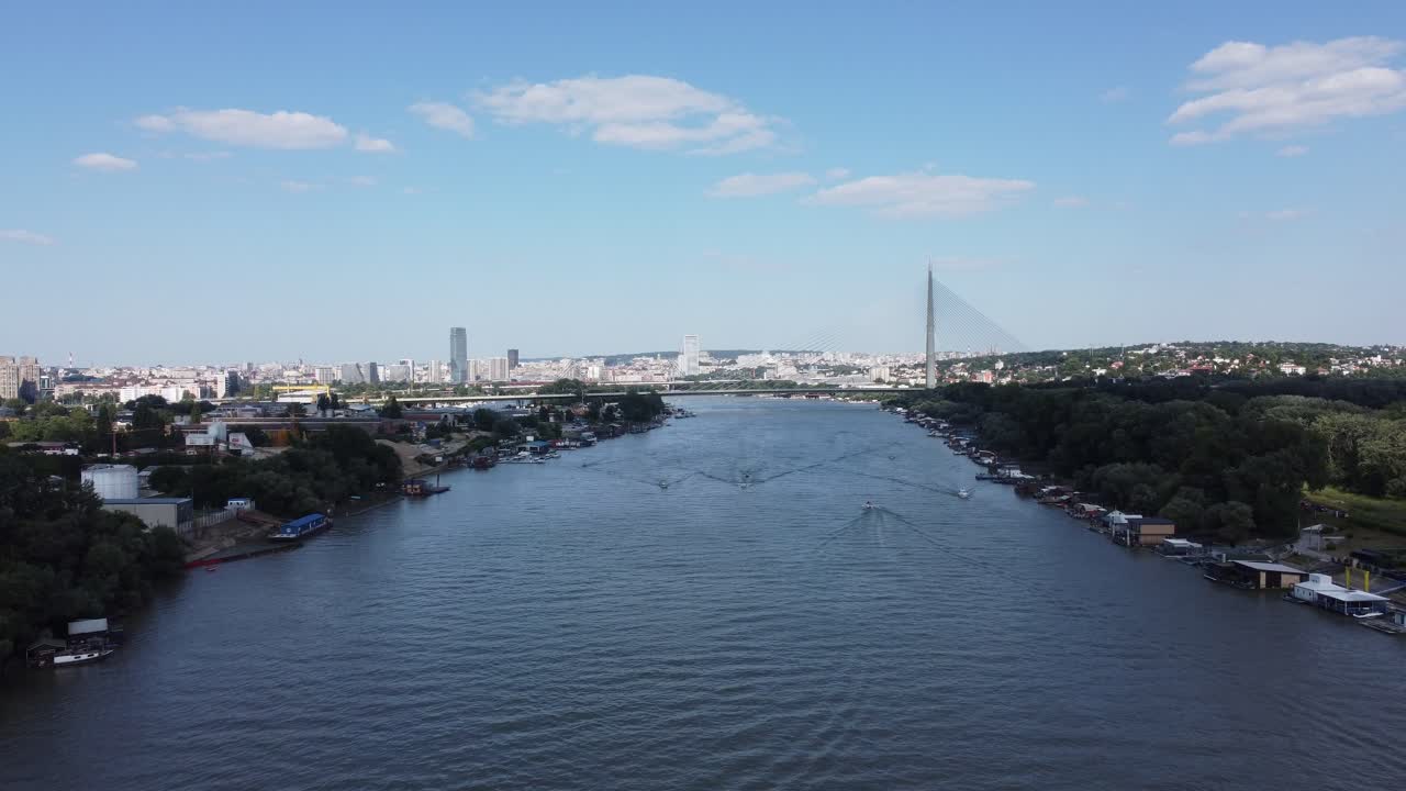 A wide river flows gracefully through a bustling cityscape, flanked by buildings, trees, and various urban structures. Boats and small docks line the riverbanks