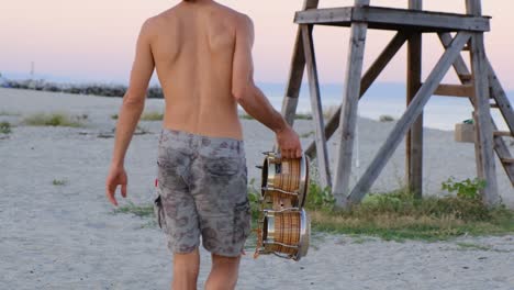 man walking half naked carrying bongo drum on a white sand beach in greece during summer