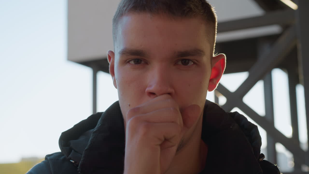 close up of sick boy outdoors coughing with open mouth under sunlight, looking unwell with slightly tired expression on face, standing near metal structure in urban environment during daylight