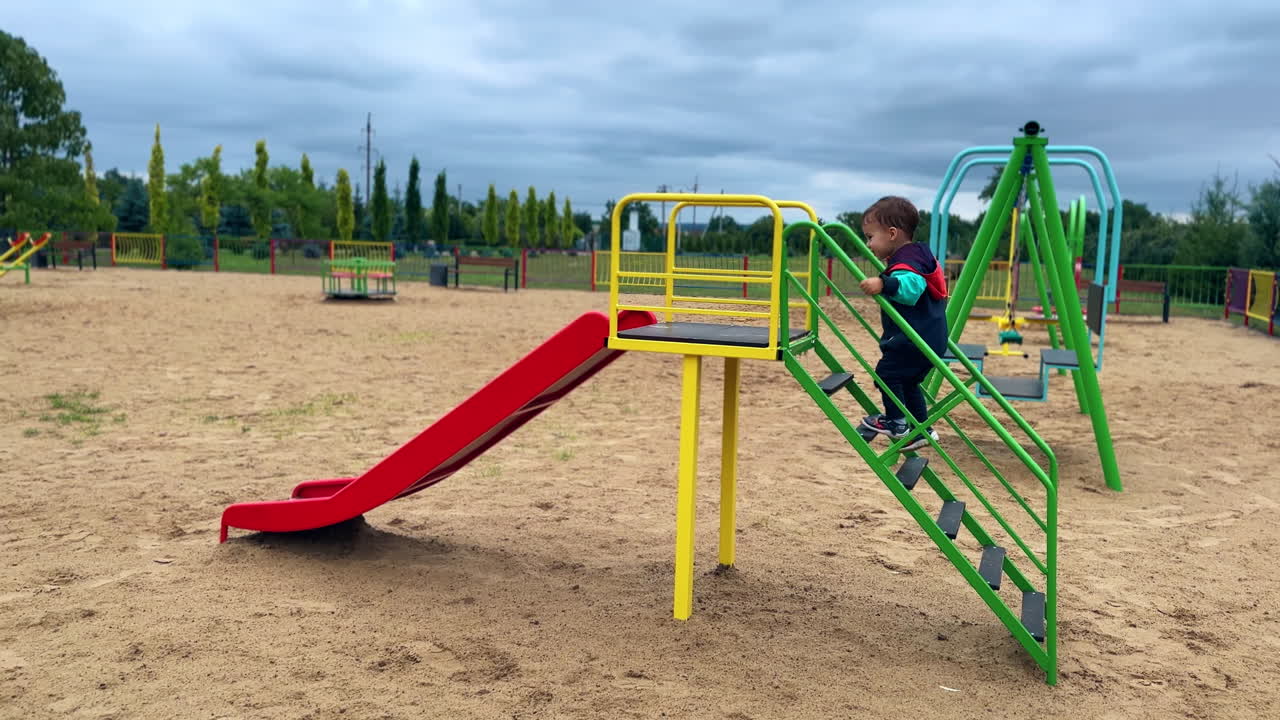 Adorable toddler runs up to the slide on the playground. Kid goes up by the stairs, sits banging feet and slides down.