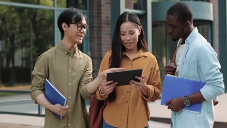 大学の近くに立って何かを議論しながらタブレットを見ている 3 人のアジアとアフリカ系アメリカ人の学生