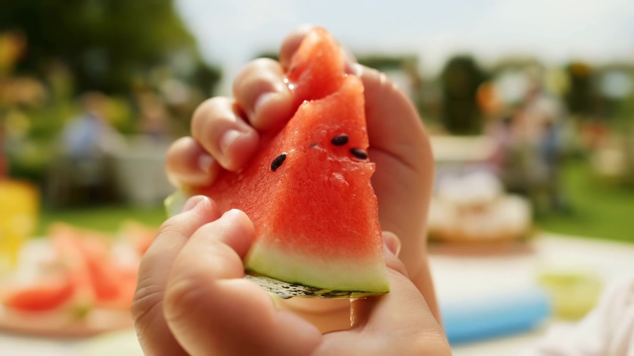Vista ravvicinata delle mani di un bambino che tengono una fetta triangolare di anguria, polpa rossa con semi neri, buccia verde chiaro, consistenza umida, luce solare intensa, spuntino estivo fresco, atmosfera allegra di un picnic all'aperto.