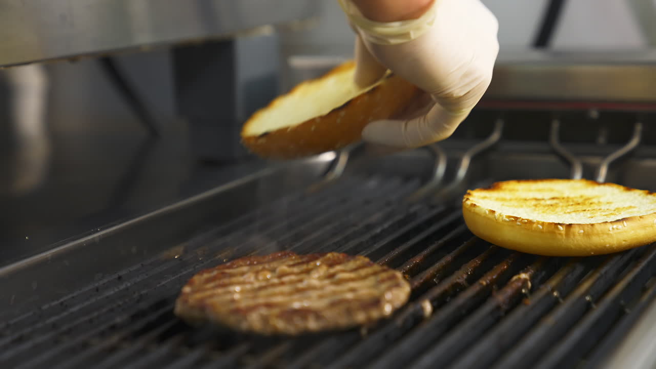 Side view of the delicious burger cutlet and two buns flipped on the grill with hand in latex gloves. Selective focus.