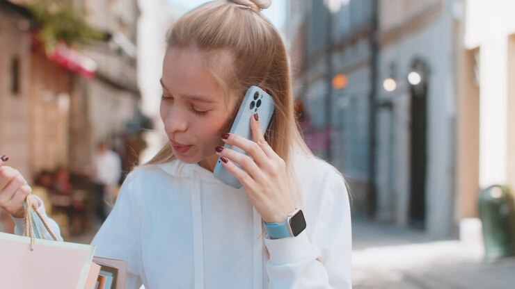 Happy young woman teenager girl talking on smartphone standing on city street holding shopping bags