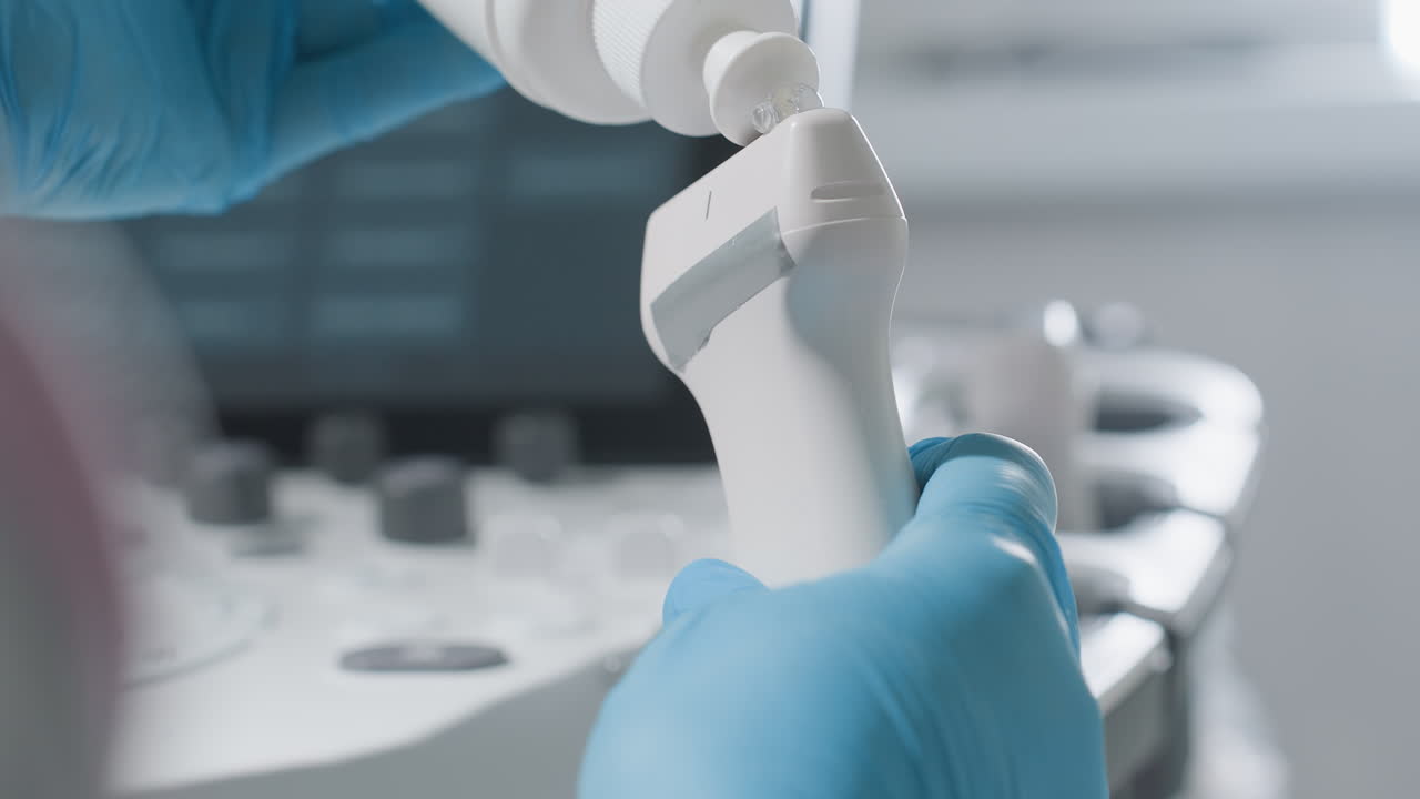 Close up of gloved hands of medical professional applying gel to ultrasound transducer while preparing diagnostic tool before patient examination in clinical setting with machine blurred