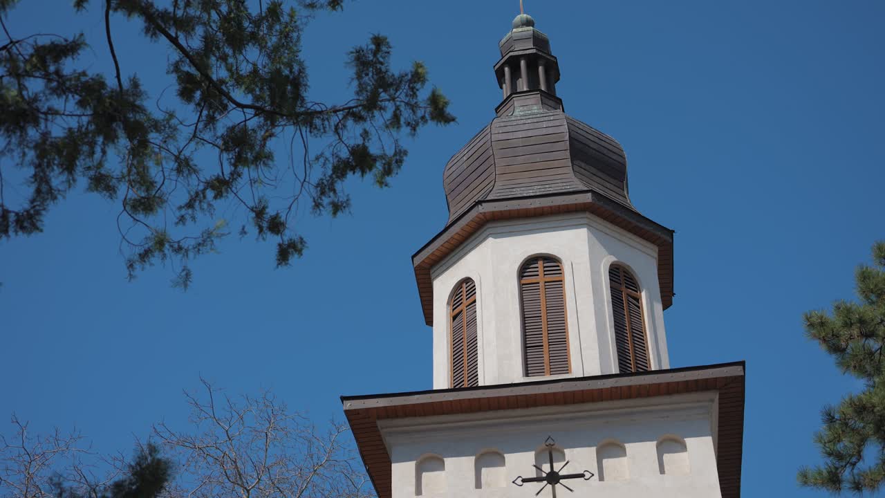 Kamerafahrt aus der Froschperspektive eines traditionellen Kirchturms unter klarem blauem Himmel, eingerahmt von umliegenden Baumästen.