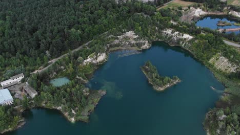 Vista-Aérea-Del-Lago-Con-Agua-Hermosa-En-Una-Cantera-Rodeada-De-Bosque-Y-Pequeña-Ciudad