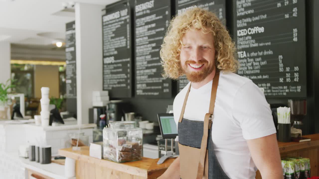 Portrait of happy caucasian male barista, smiling with arms crossed behind counter in cafe