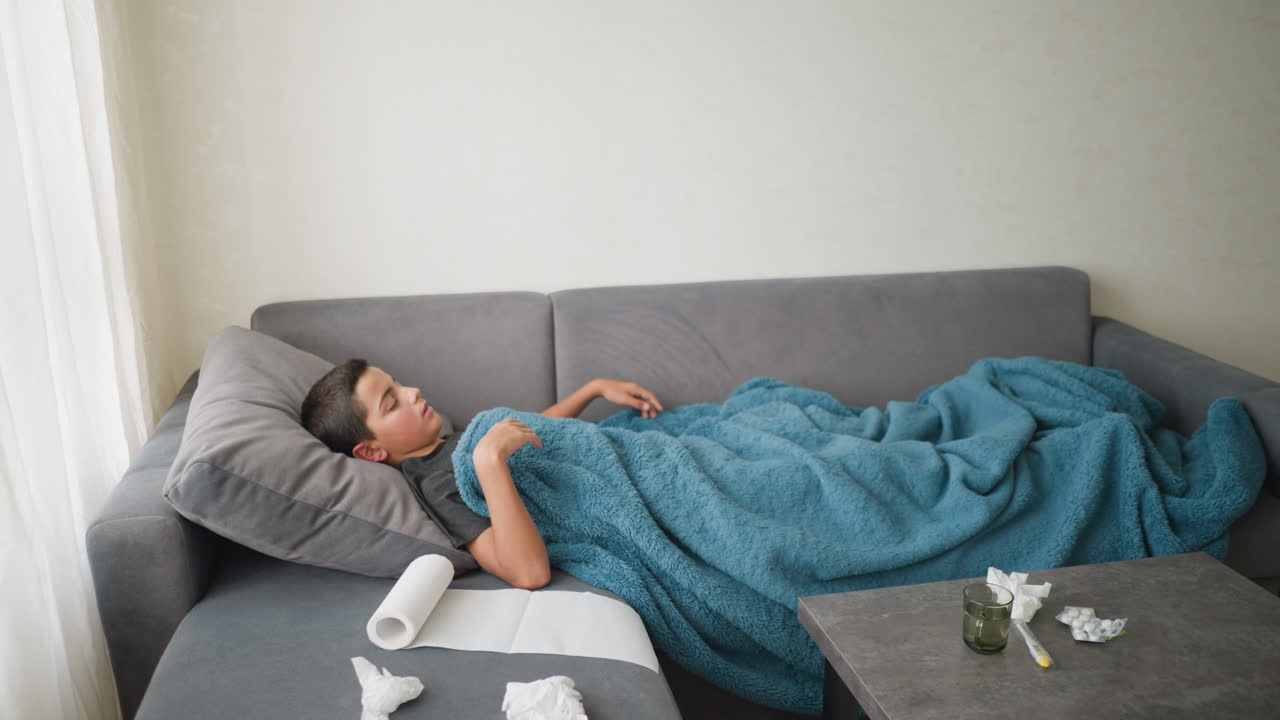Stanza in disordine con bambino sdraiato sul divano che si sistema per stare comodo, termometro per medicinali e fazzoletti usati sul tavolo, a indicare malattia e riposo, ambiente tranquillo ma disordinato in un contesto domestico