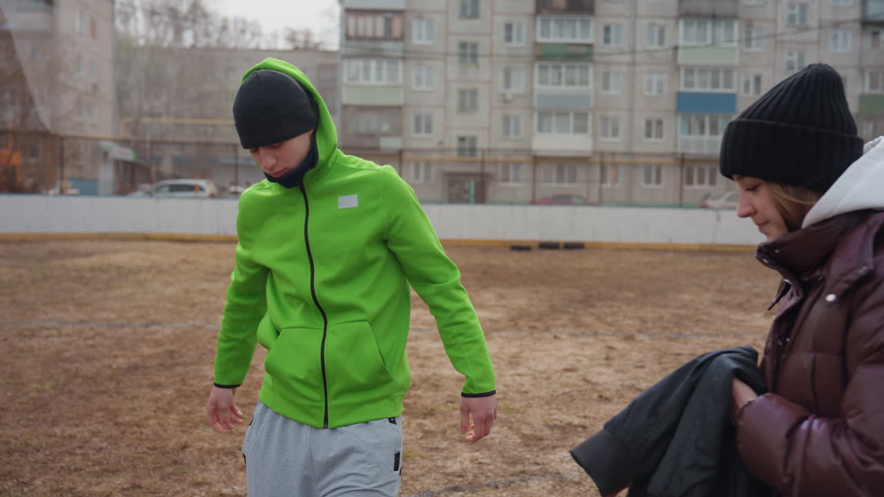 Белый подросток снимает куртку, пока друг наблюдает за ним на фоне городского пейзажа, холодной многоквартирной застройки, в шапке и пуховике, крупный план рук, обменивающихся одеждой, приглушенный зимний свет, искренние эмоции и доверие