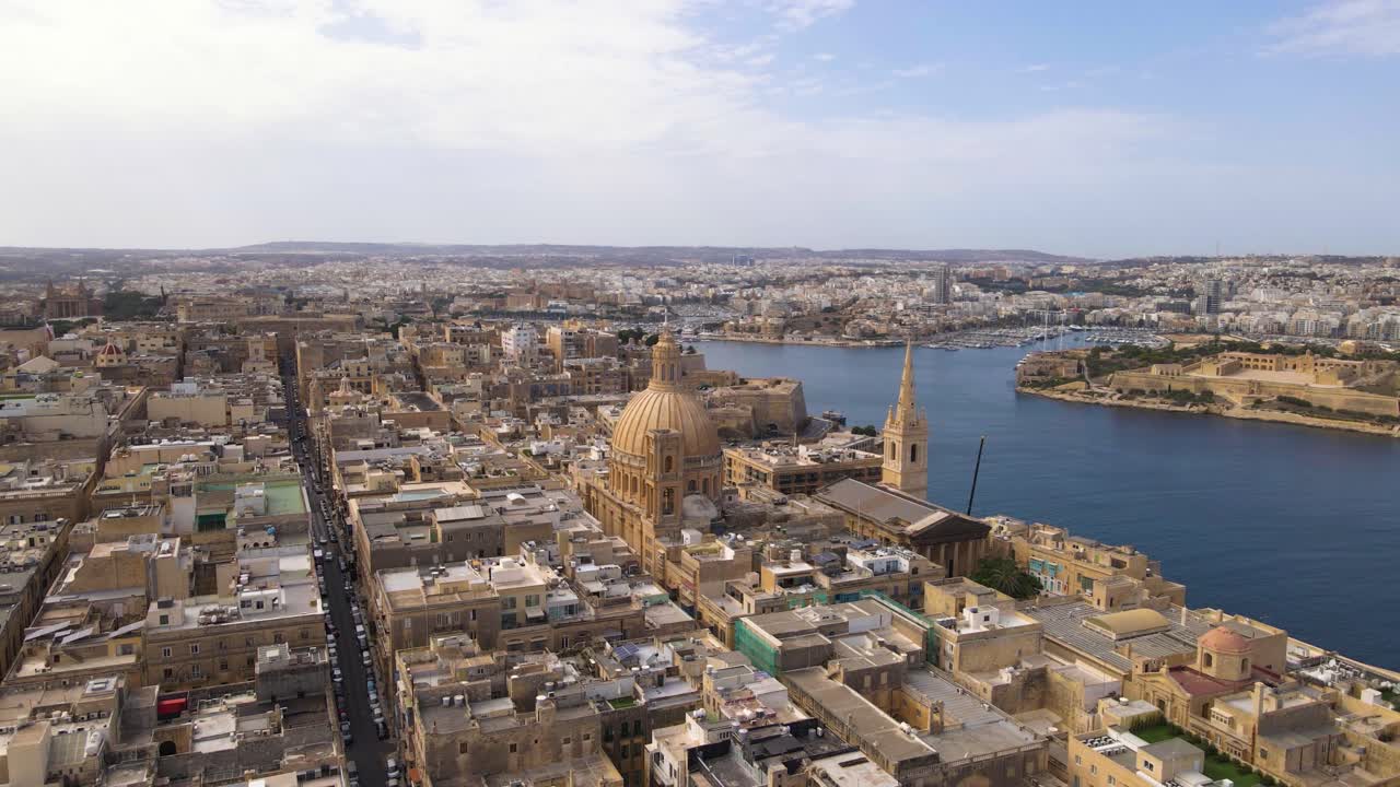Spettacolare ampia prospettiva aerea di La Valletta che mostra la maestosa cupola dorata della Chiesa Carmelitana che si erge sopra il denso paesaggio urbano di calcare, con l'ampio Grand Harbour e il porto turistico oltre.