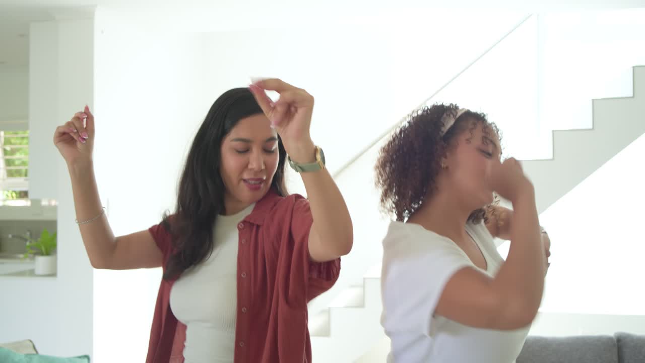 Diverse female friends leaning back-to-back on gray sofa, dancing, spinning, moving to coffee table