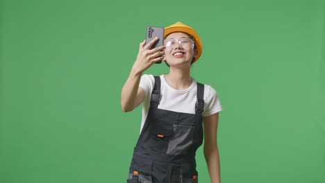 안경과 안전 헬을 착용 한 아시아 여성 노동자가 녹색 화면 배경 스튜디오에 서있는 동안 스마트폰에서 비디오 통화를 하고 손을 흔들고 있습니다.