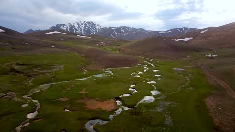 aerial green highland with snowy mountains landscape and beautiful stream reflects the cloudy sky in spring day time in kerman iran