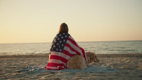 blonde girl sitting on a sunny beach with her dog wrapped in an american flag on a sunny beach in the morning