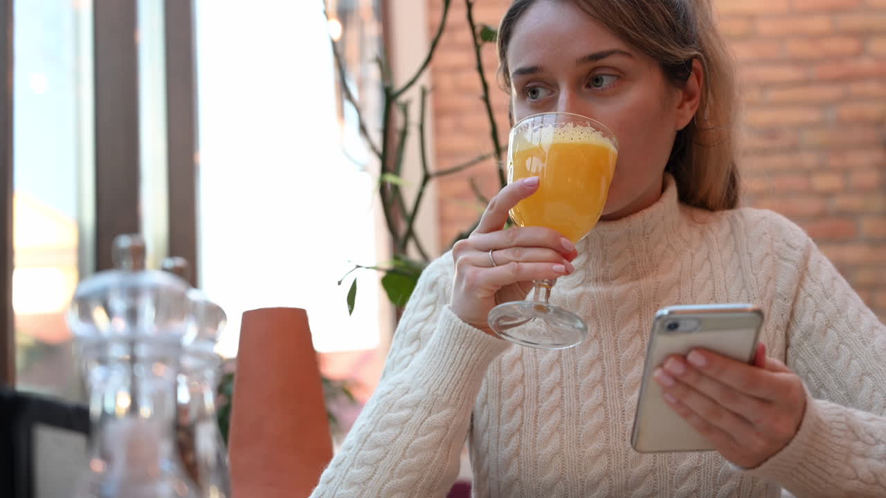 Close up of a woman in a white turtleneck scrolling on her phone and drinking orange juice in a restaurant