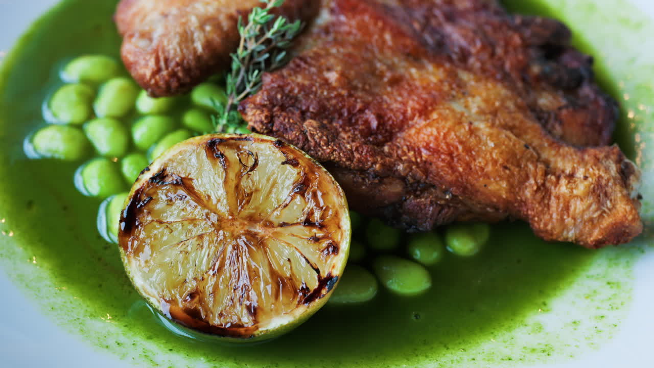 Close-up of a gourmet roasted chicken thigh served with edamame beans, a drizzle of herb-infused green sauce, and a charred lime half
