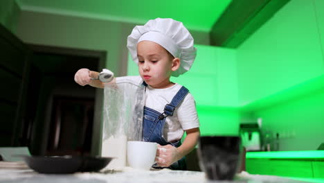 kid in white chef cap holds a long container and gets flour from it. toddler boy puts wheat flour to the plate. low angle view.