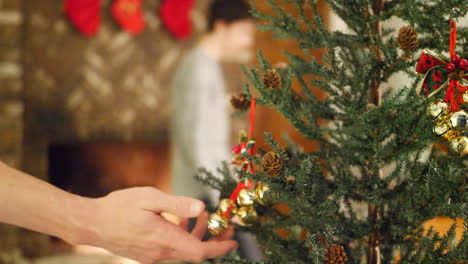 a person hanging a jinglebell ornament onto a christmas tree with a fire and stocking over the fireplace for the holidays