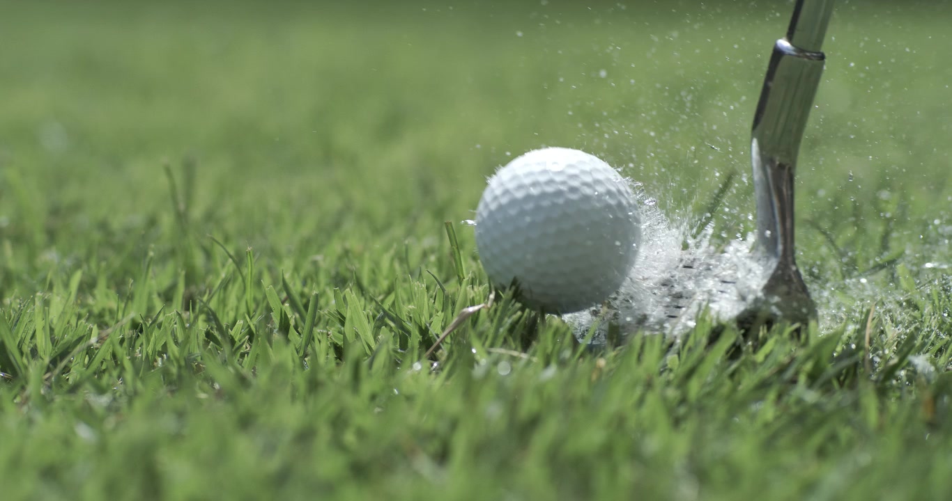 Golfclub raakt een witte golfbal in super slow motion. Druppels ochtenddauw en grasdeeltjes stijgen op in de lucht na de impact. Gefilmd met een super slow motion camera 1000 fps