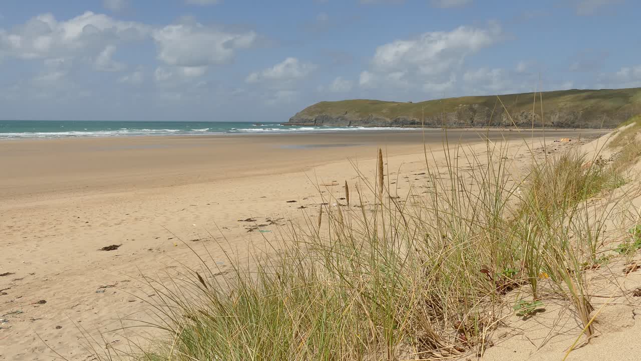 Peaceful view of golden sand beach in Cornwall with grassy dunes, turquoise waves, and rugged cliffs under blue sky, showing natural coastal beauty and calm seaside atmosphere of England
