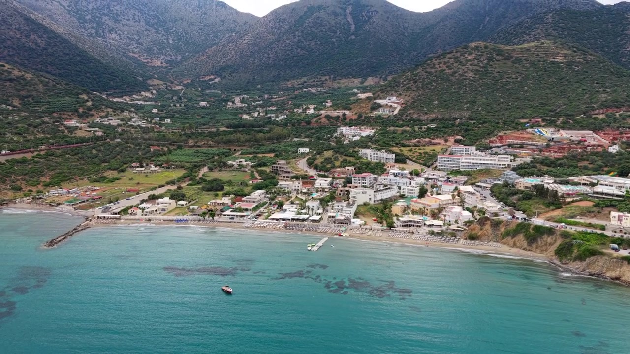 Vista aérea de drone de uma baía de resort e praia de areia com guarda-sóis, uma cidade em um vale verde abaixo de montanhas íngremes, Bali, Grécia