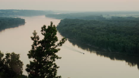 distant motorboat glides across wide river lined with lush mangrove forest under soft dusk sky as ripples trail behind boat cutting through reflective water surface