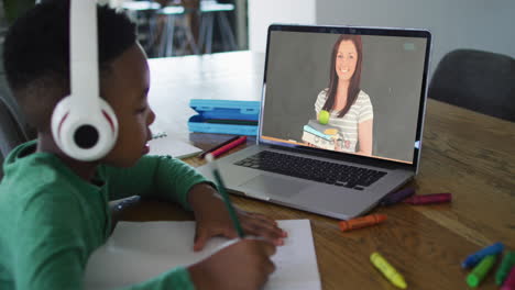 african american boy wearing headphones having a video call on laptop while doing homework at home