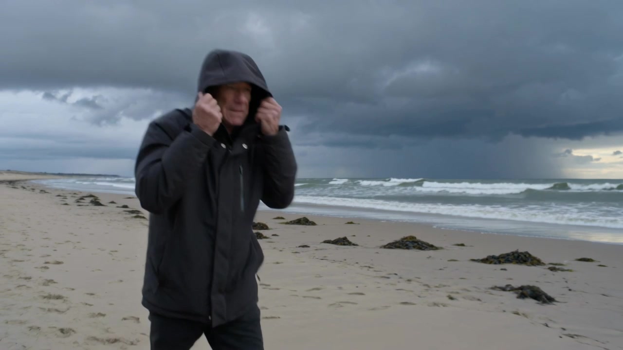 Walking man pulling dark hooded jacket up on sandy beach, shielding from approaching storm and wind