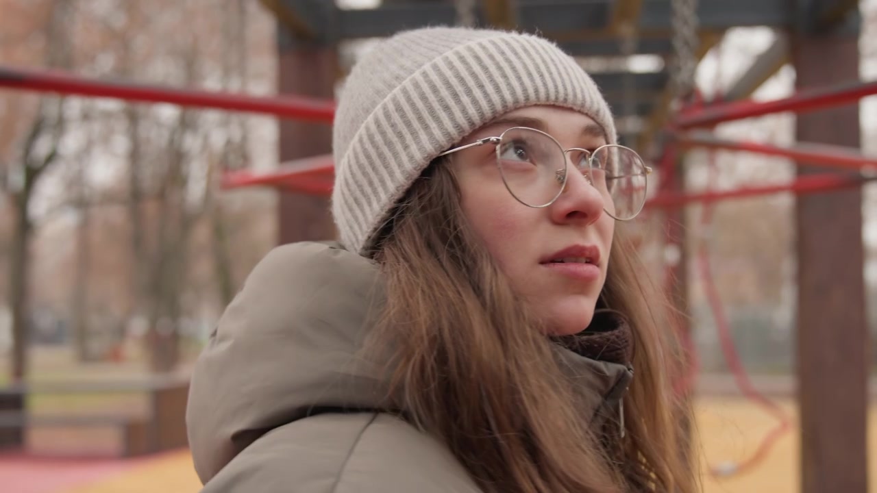Giovane donna bianca su una struttura di corde del parco giochi, con berretto di lana e occhiali, piumino, sullo sfondo alberi autunnali. Ritratti ravvicinati che mostrano uno sguardo pensieroso e una curiosità silenziosa, atmosfera cinematografica lenta, urbana.