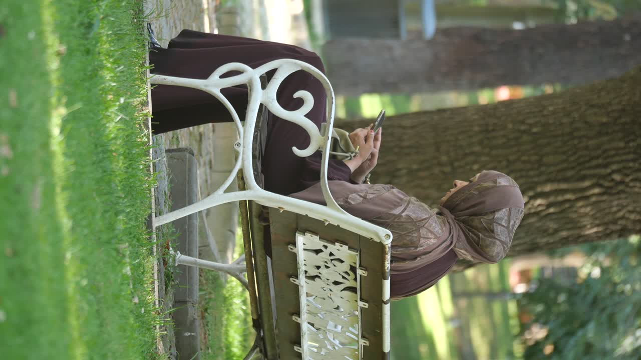 Woman in hijab sitting on park bench using phone