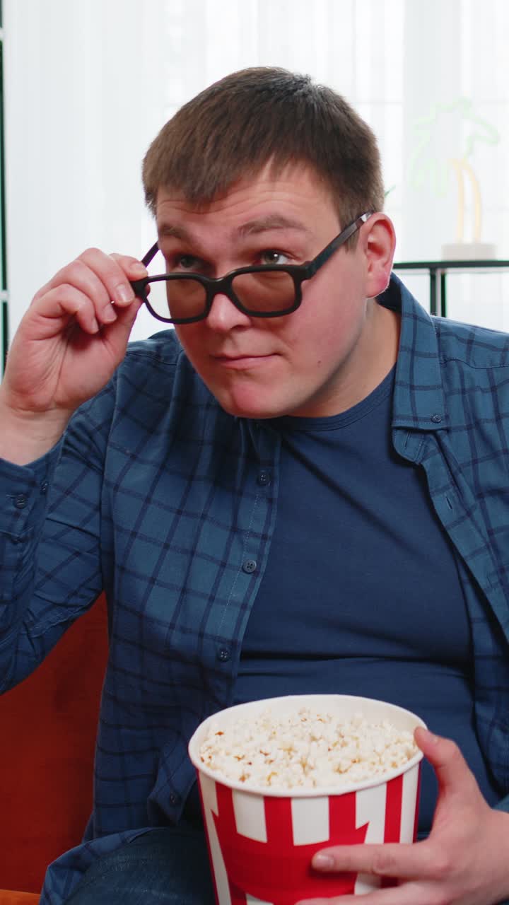 Caucasian man in 3d glasses sitting on couch eating popcorn snacks and watching interesting tv film