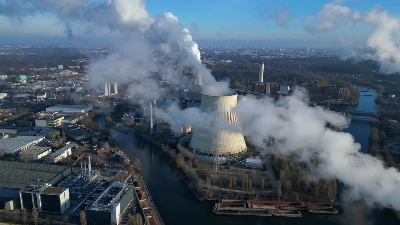 Aerial view of a thermal power plant emitting smoke on a sunny day, with a river and a city in the background. Wonderful aerial view flight panorama overview drone
