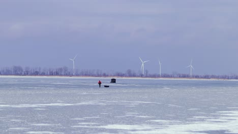 Pescadores-Despertando-En-El-Lago-Congelado,-Tirando-Trineos-De-Hielo,-Pesca-En-Hielo,-Invierno-Frío-Al-Aire-Libre,-Carpa-De-Pesca-En-El-Lago-Congelado,-Mid-Shot-Canada-Amplia-Vista-Del-Paisaje,-Molino-De-Viento,-Energía-Renovable