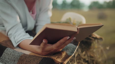 close-up of a woman hand with a bangle turning to a new page on a blanket outdoors, enveloped in a golden glow, with a blurry view of a hat by the side