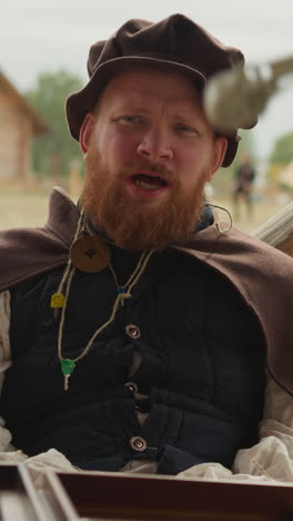 red-bearded squire talks to woman sitting at table in courtyard in medieval village. female hand spins pendant on rope in circle on blurred background closeup