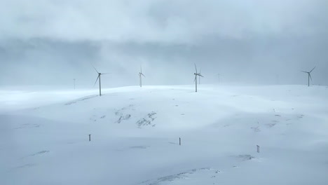 Schwenkbare-Luftaufnahme-Eines-Windparks-In-Einem-Schneesturm-In-Nordnorwegen-Mit-Sich-Drehenden-Windkraftanlagen-In-Einer-Hügeligen-Winterlandschaft-In-Einem-Schneesturm