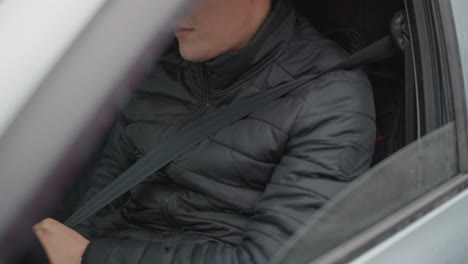 Window view of guy seated in driver seat pulling seat belt strap across torso beside reflective window showing pedestrians passing by highlighting safety gesture and motion during commute
