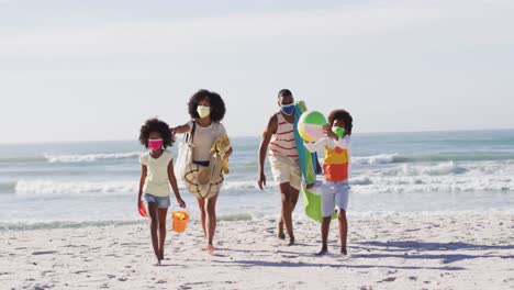 African-american-parents-and-their-children-wearing-face-masks-carrying-beach-equipment-on-the-beach